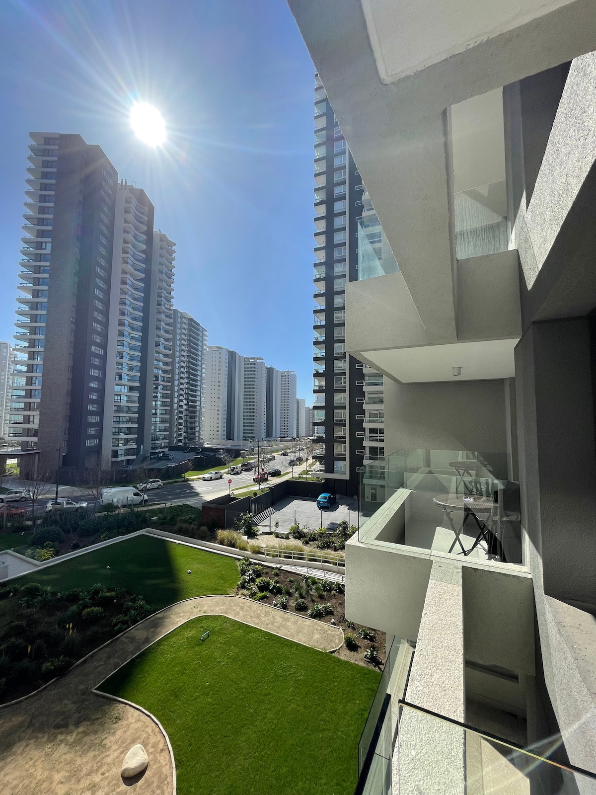A modern balcony offers a view of the surrounding high-rise buildings and landscaped gardens. Bright sunlight illuminates the outdoor space, which features a small table and chair. Green grass and pathways are visible below, contributing to the overall urban yet natural atmosphere.