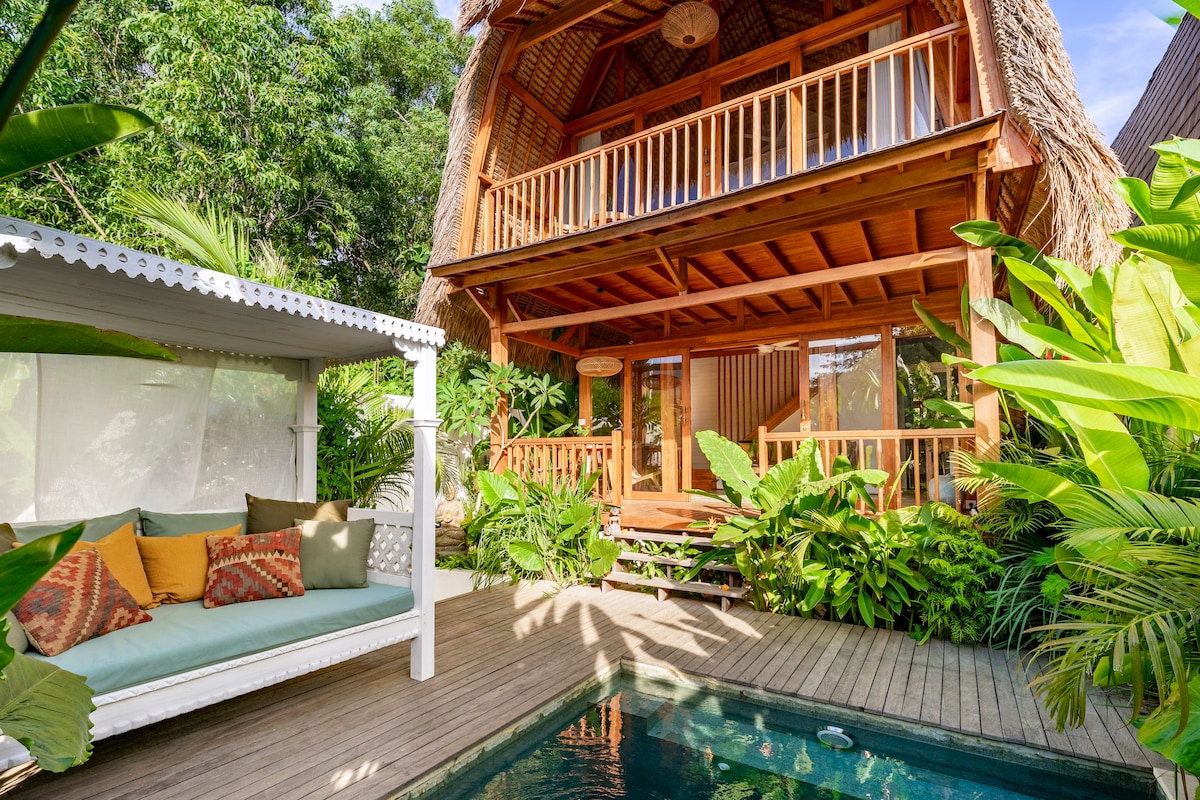 A traditional Balinese building with a thatched roof is surrounded by lush greenery. A private pool is situated in front, complemented by a comfortable lounge area with cushions. The upper balcony features wooden railings, offering a view of the garden below.