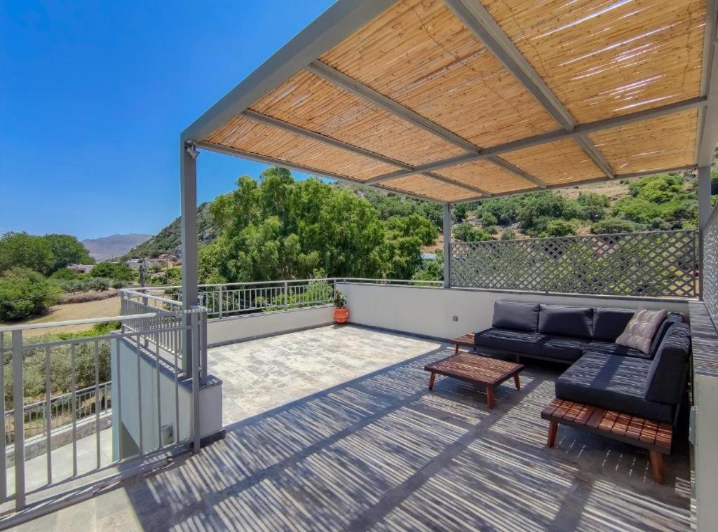 A spacious terrace features a covered seating area with a comfortable L-shaped sofa and a wooden coffee table. Natural light filters through the bamboo roof, casting gentle shadows on the floor, while scenic views of the surrounding hills are visible in the background.