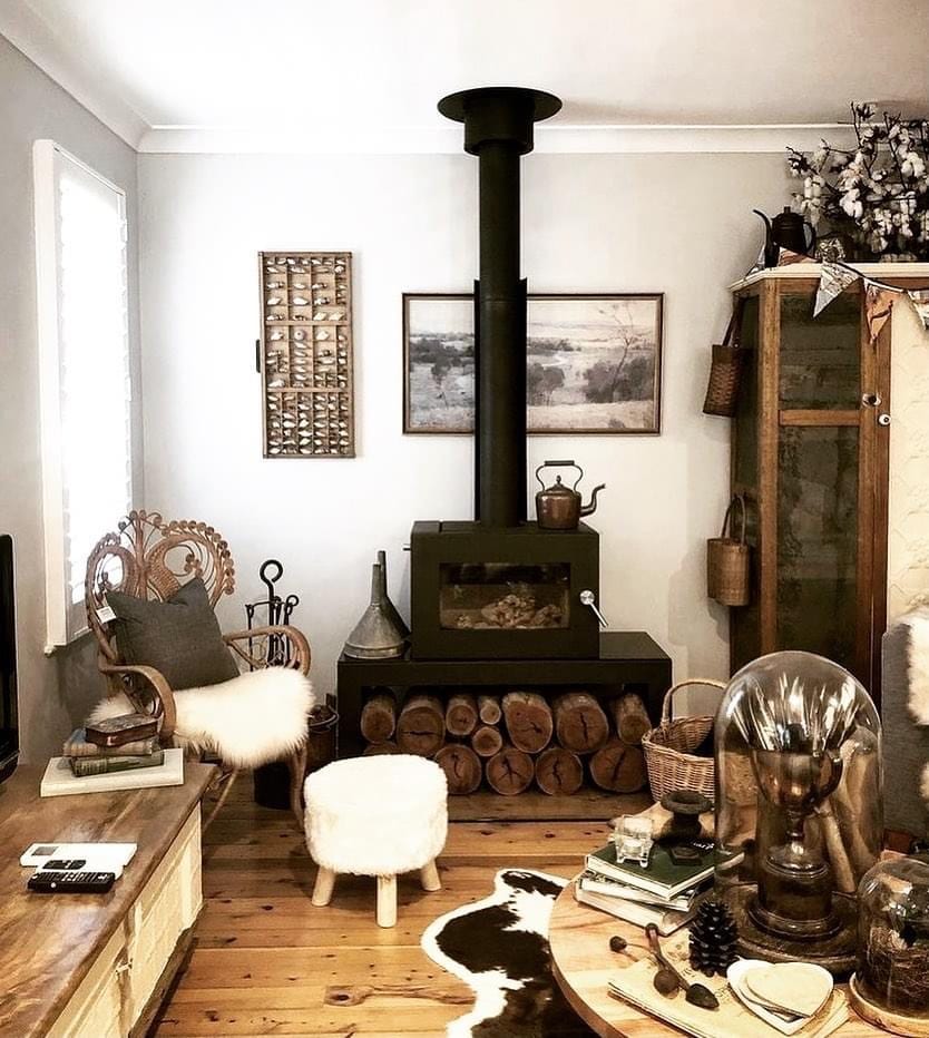 A vintage-style living space features a wood-burning fireplace set against a backdrop of light gray walls. A plush chair and a small stool are positioned nearby. The floor is adorned with a decorative rug, while shelves display various unique items and plants.