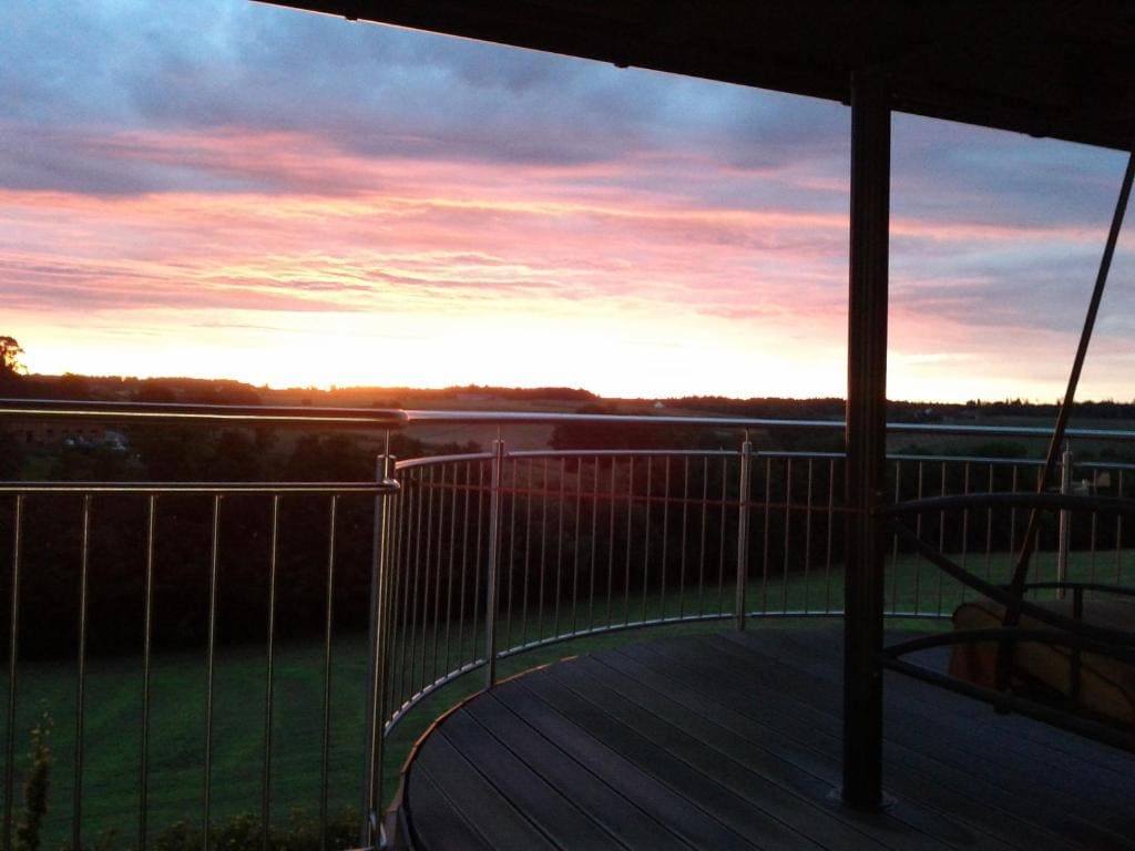 A balcony offers a panoramic view of the horizon at sunset, with vibrant hues of orange and pink illuminating the sky. The railing surrounds a spacious deck that overlooks a lush green landscape, creating a serene and picturesque setting.