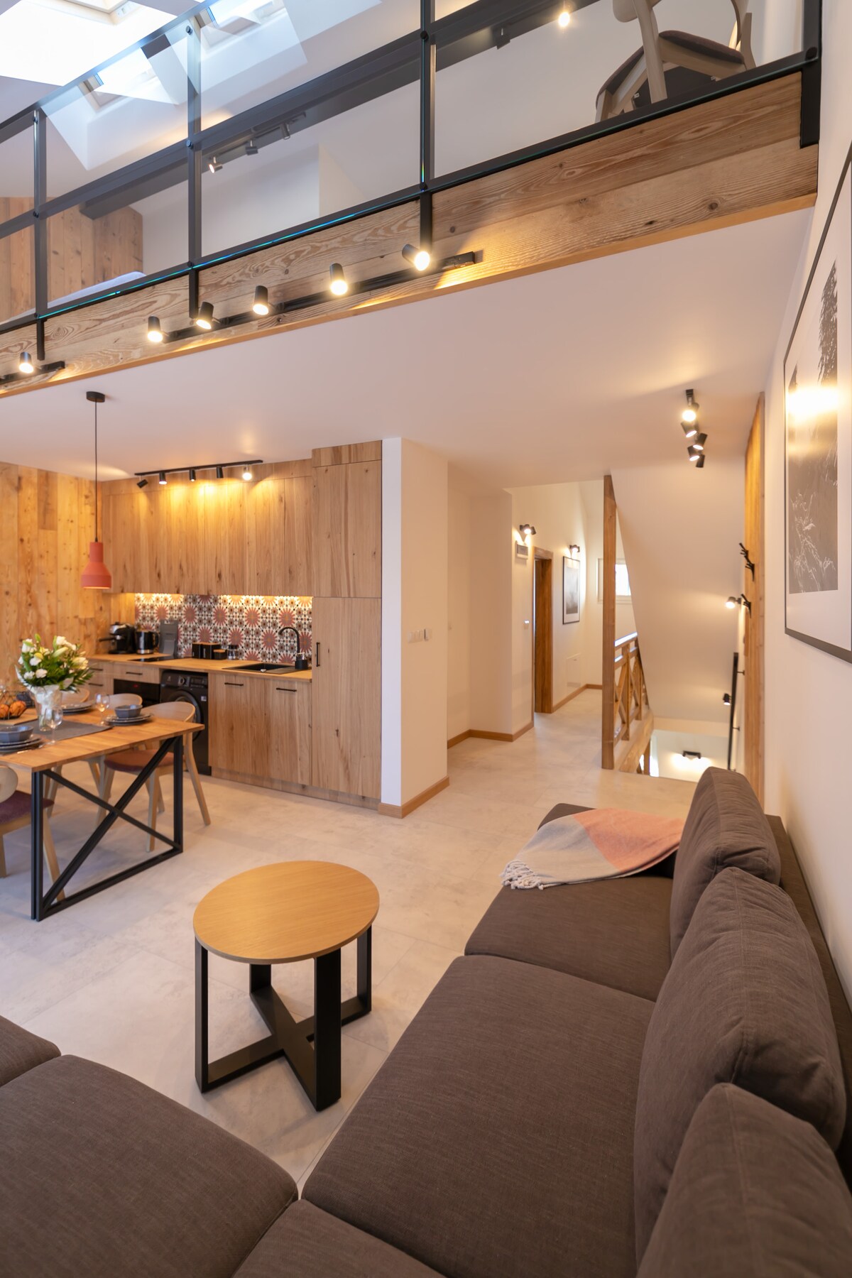 A modern living area includes a comfortable gray sofa, a round side table, and a dining space with wooden accents. The kitchen features a decorative backsplash, highlighting an inviting atmosphere. Soft lighting enhances the warm tones, while a staircase leads to an upper level.