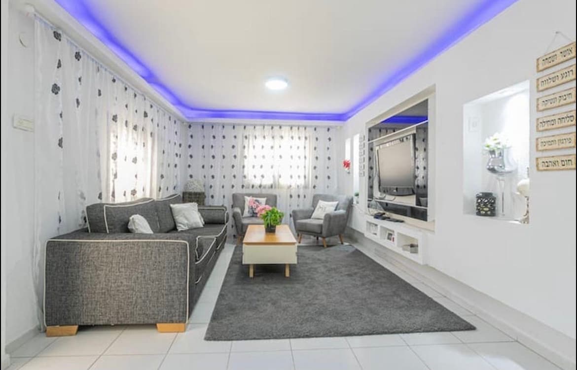 A comfortable living area is highlighted by a soft gray sofa and two accent chairs. A decorative coffee table sits on a plush rug, and natural light enters through the patterned curtains. Soft lighting is provided by a ceiling fixture and ambient blue accents.