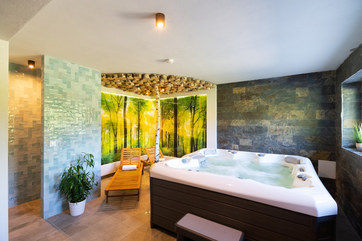 A serene spa area is presented with a white hot tub surrounded by natural stone walls. A vibrant mural depicting a forest adds a calming ambiance. A wooden lounge chair is placed beside the tub, with indoor plants providing a touch of greenery.
