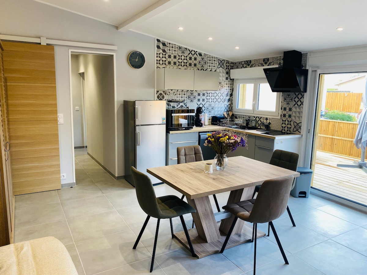 A modern kitchen and dining area feature a wooden table surrounded by four upholstered chairs. Clean lines are accentuated by stylish tile and cabinetry, while a large window lets in natural light. Kitchen appliances, including a refrigerator and coffee maker, are conveniently arranged.