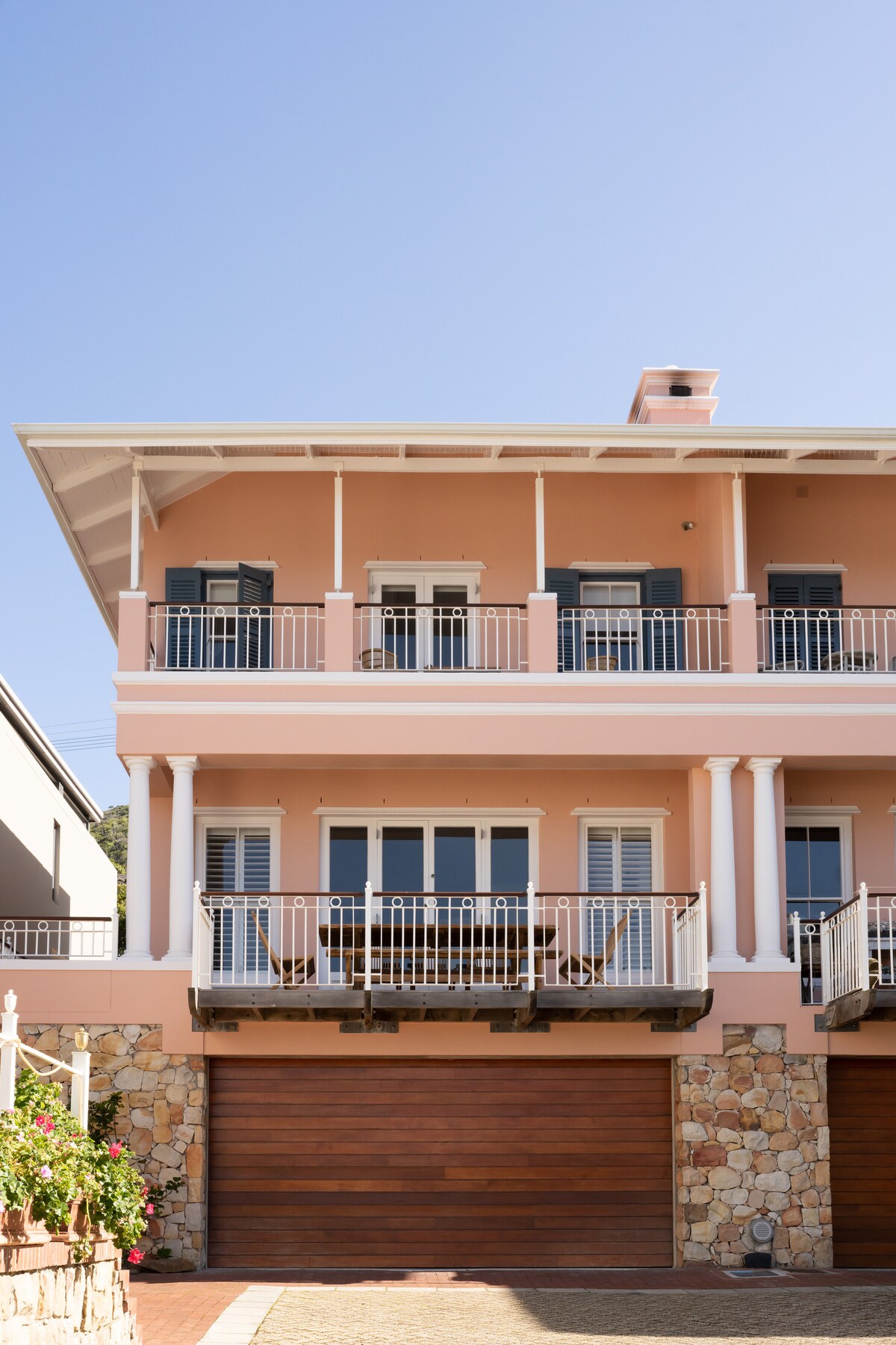 The exterior of a three-story house is presented, showcasing a soft pink façade and decorative balconies adorned with white railings. Detailed stonework supports the lower level, while large windows allow natural light into the home. A serene blue sky serves as a backdrop.