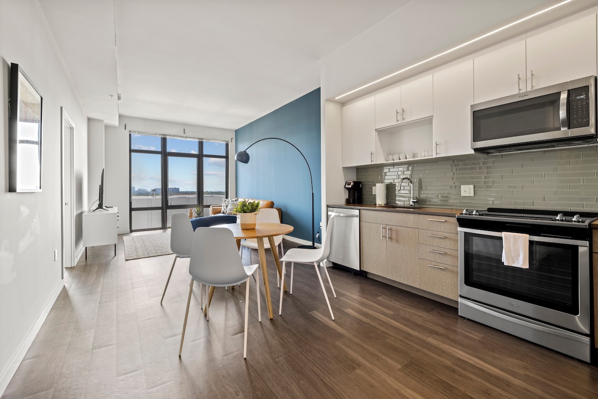 A modern kitchen and dining area are showcased, featuring light wood flooring. A round dining table is surrounded by four white chairs, while stainless steel appliances and a stylish backsplash complete the kitchen. Large windows offer natural light and views of the outside.