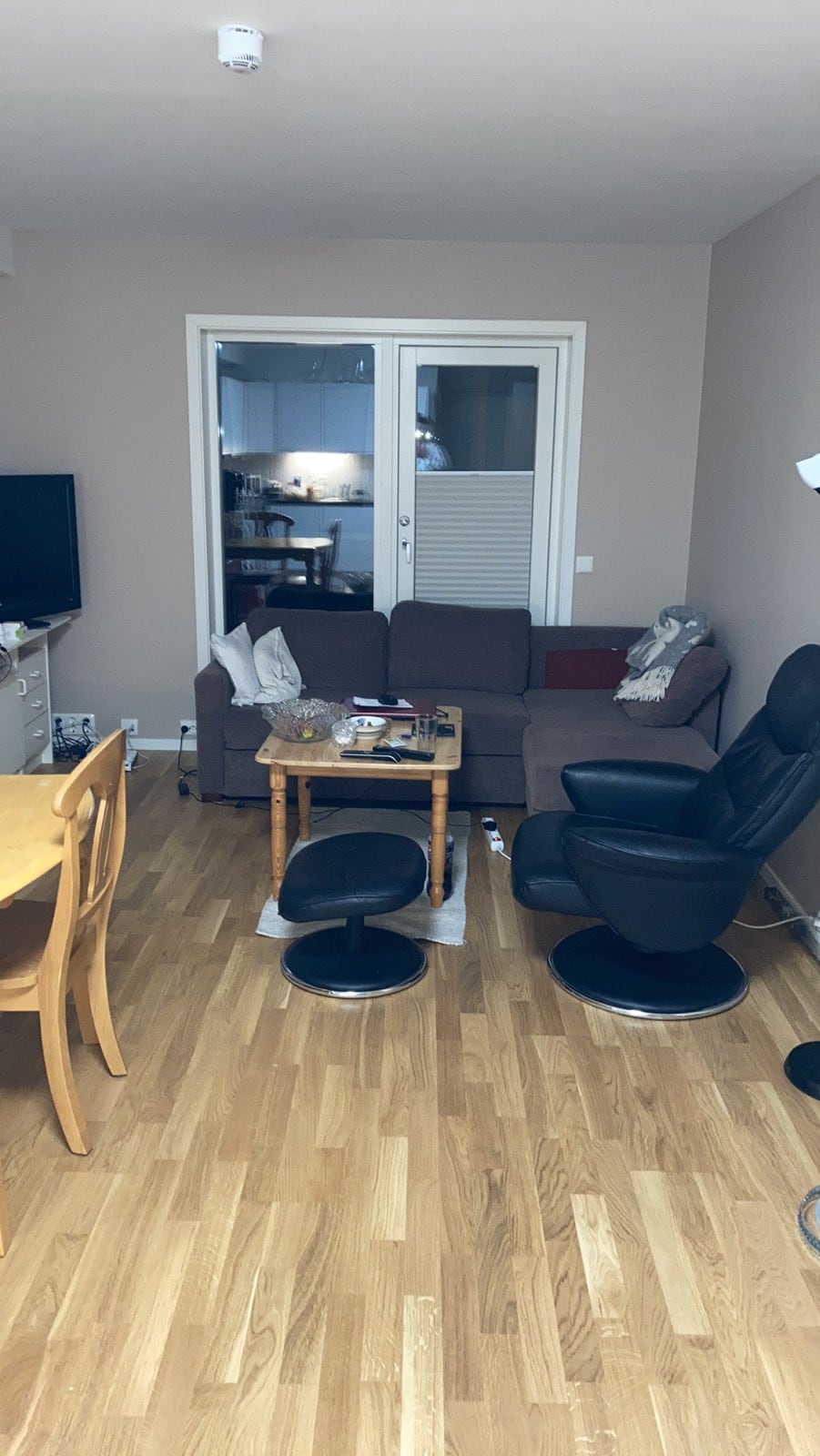 A cozy living area features a brown sofa facing a wooden coffee table. A black swivel chair is positioned nearby, and a dining table with wooden chairs is visible. Light is seen from a window leading to the kitchen area, highlighting the wood flooring.
