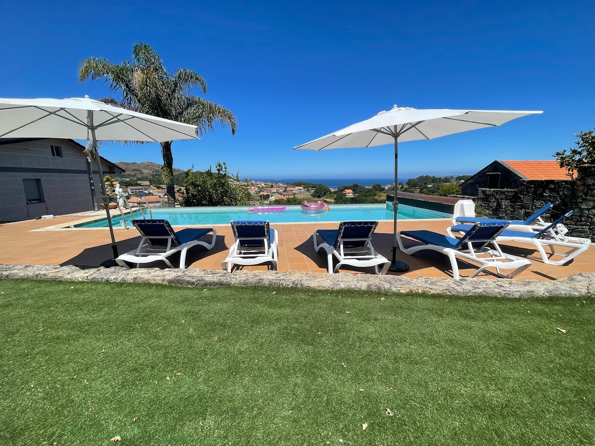 A private pool area is outlined by sun loungers under large umbrellas. Lush greenery surrounds the space, with views of the ocean and nearby landscape in the background. The patio features a smooth wooden deck, providing a serene setting for relaxation.