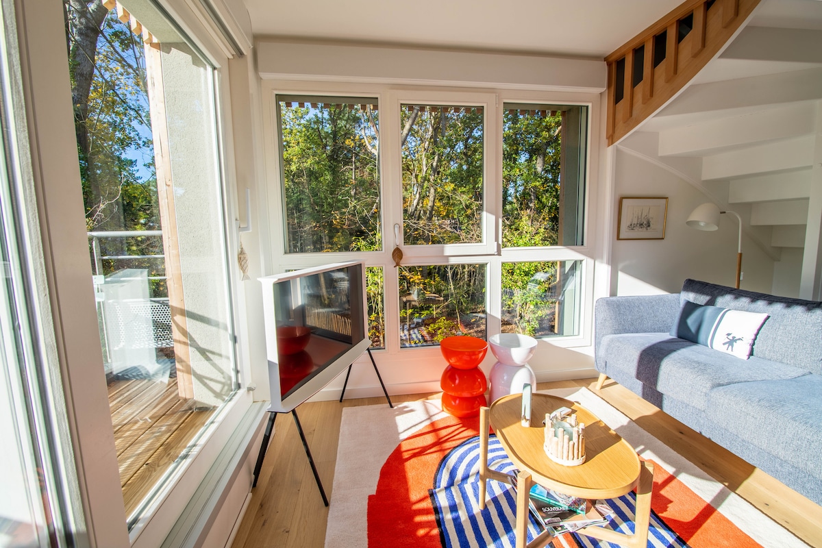 A bright living area is illuminated by natural light streaming through large windows. A modern sofa in a calm shade contrasts with an orange accent chair. A decorative area rug is placed under a wooden coffee table, creating a cozy focal point in the room.