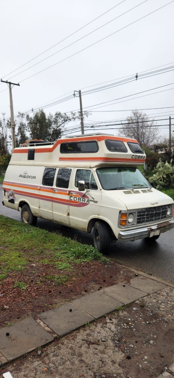 A vintage camper van is parked alongside a street, showcasing its distinctive orange and white exterior design. The van features large windows on the roof, providing an elevated view. Surrounding greenery is present, complementing the vehicle's rustic appearance.