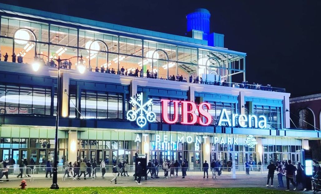 The UBS Arena at night features a modern facade with large glass windows, showcasing illuminated spaces inside. People can be seen entering the venue, while a vibrant sign identifies the arena prominently. Surrounding greenery and lighting enhance the atmosphere.