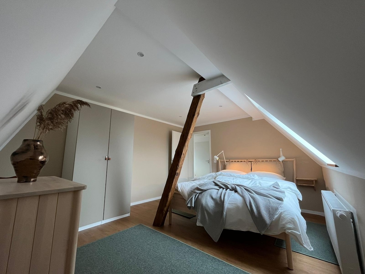 A cozy bedroom features a spacious bed with light bedding, positioned beneath a sloped ceiling. Natural light filters through a small window, highlighting wooden beams and a stylish vase on a dresser. A soft rug adds warmth to the wooden floor.