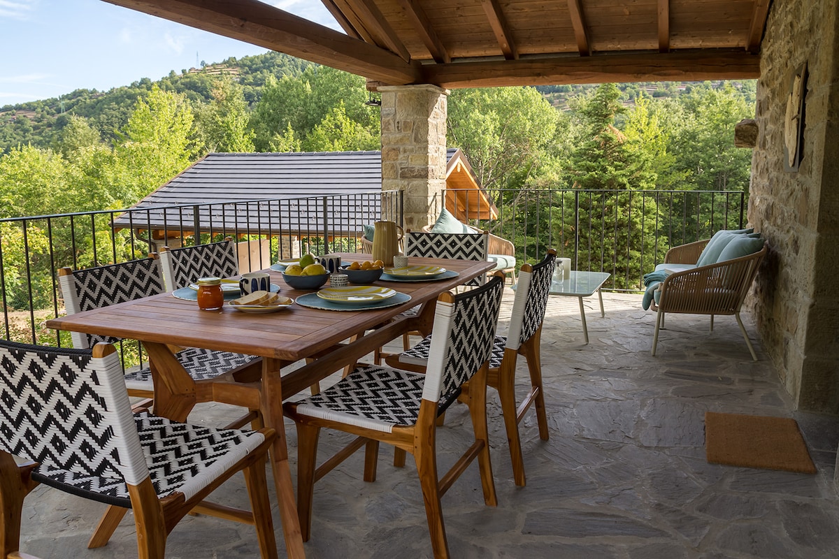 A shaded outdoor dining area features a wooden table surrounded by patterned chairs. Plates and cutlery are set, creating a welcoming space for meals. A cozy seating area is visible, with soft furnishings overlooking the lush green forest, inviting relaxation amidst nature.