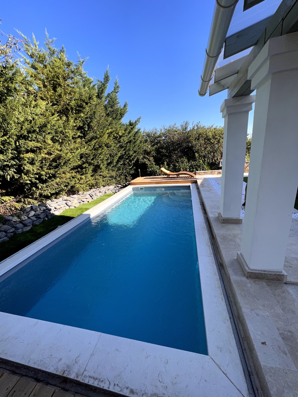 A serene backyard pool is lined with elegant stone decking. The clear blue water reflects the bright sky, while green foliage provides a natural backdrop. Sun loungers are positioned nearby, creating a peaceful retreat for relaxation.