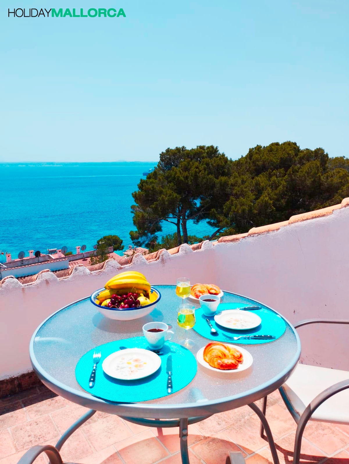 A round dining table is set with plates and utensils, featuring fresh fruit, pastries, and beverages. A clear view of the sea and trees is captured in the background, complemented by a bright blue sky. The scene reflects a serene outdoor dining experience.