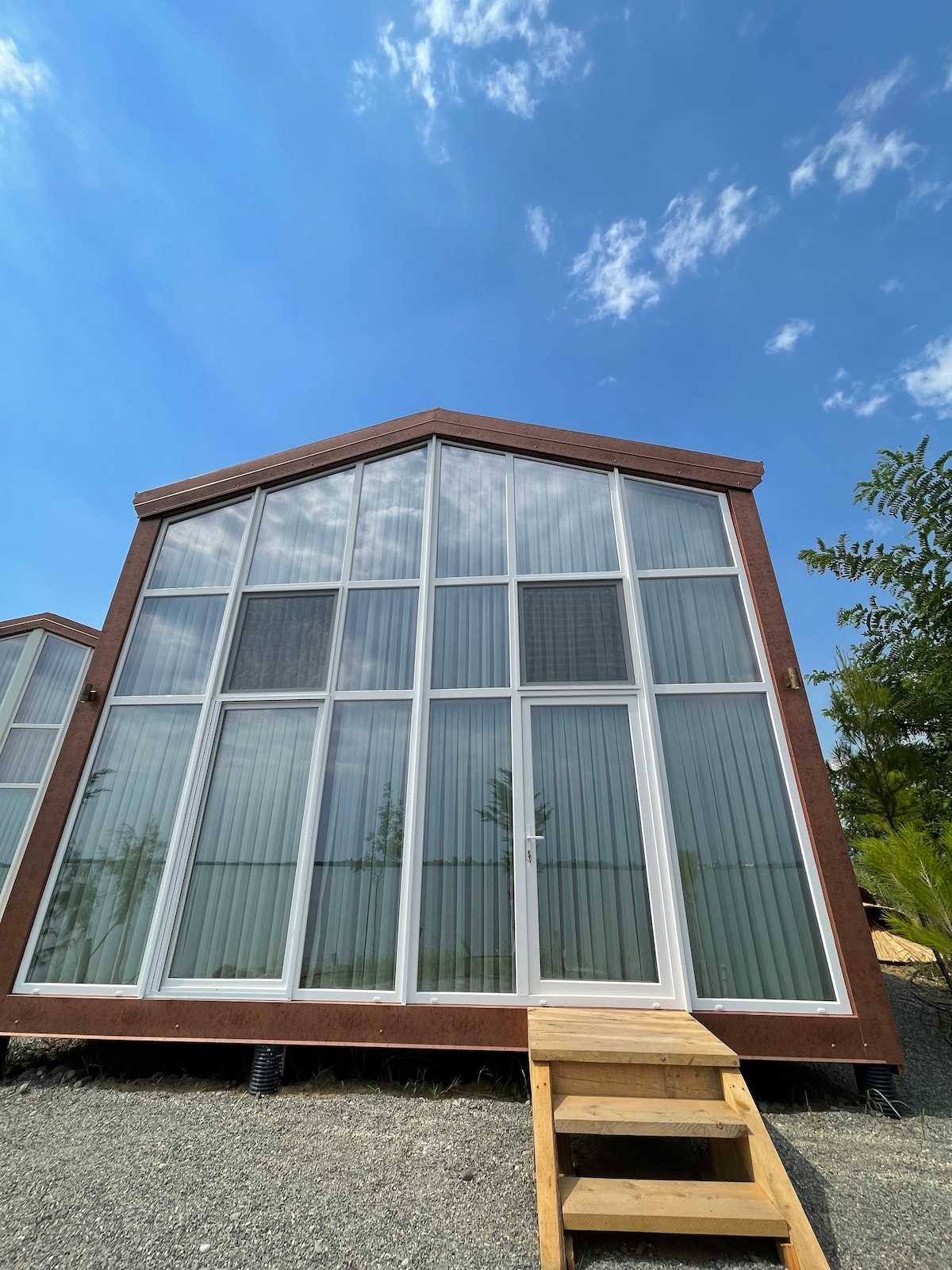 The exterior of the accommodation is showcased, featuring a modern design with a sloped roof. Large windows dominate the front, allowing ample natural light. A wooden staircase leads up to the entrance, surrounded by a gravel area and green foliage in the background.