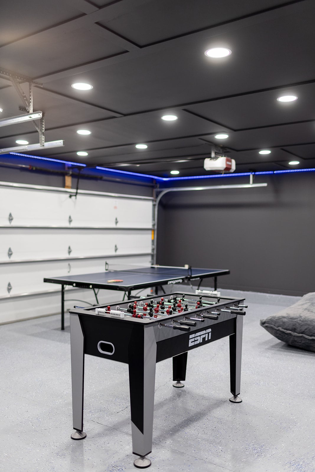 A game room is displayed featuring a foosball table and a ping pong table. The space is illuminated by recessed lighting and includes a projection screen on the ceiling. A soft seating area provides a cozy spot for relaxation.