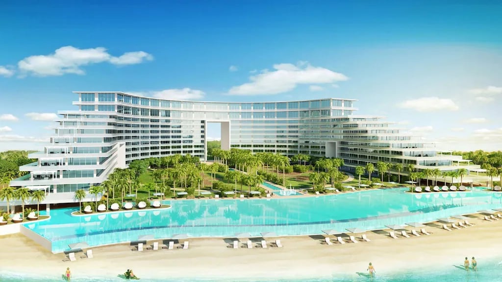 A modern beachfront property is set against a clear blue sky, featuring multiple levels and large windows. An expansive infinity pool extends towards the beach, surrounded by palm trees and lounge chairs. The area is complemented by well-maintained gardens and a serene sandy shore.