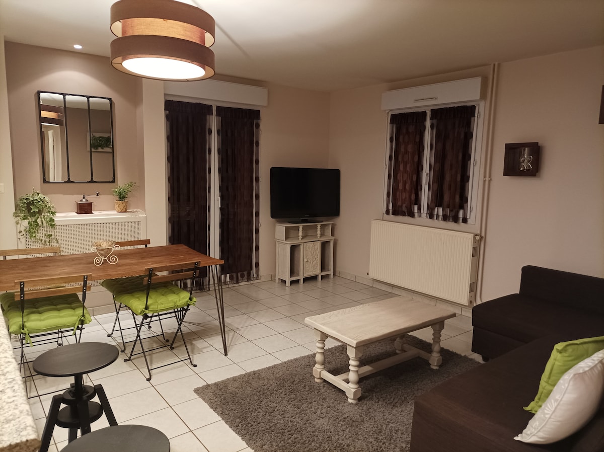 A cozy living area features a wooden dining table surrounded by chairs with green cushions. A comfortable sofa faces a mounted television, and a coffee table rests on a soft area rug. Large windows with dark curtains allow natural light to fill the space.