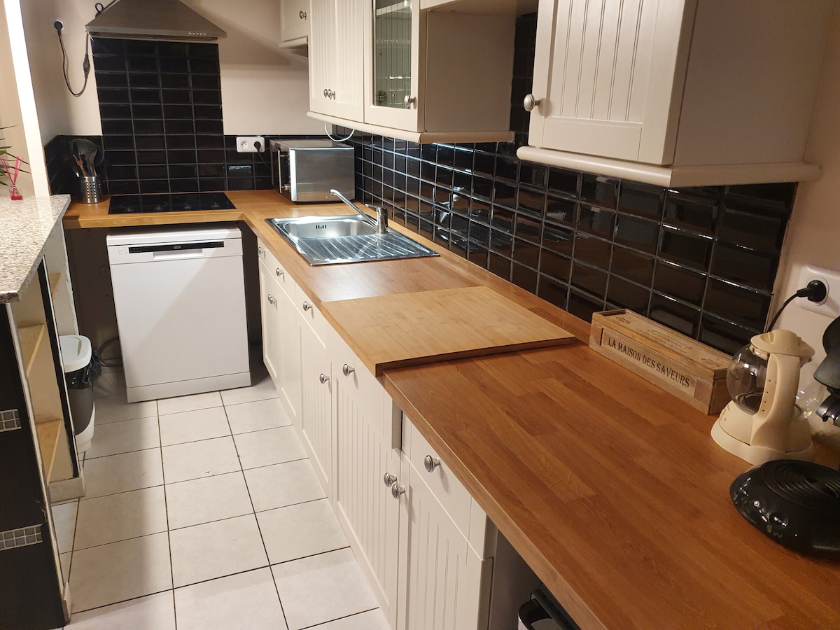 A functional kitchen is presented with wooden countertops and white cabinetry. An integrated dishwasher and a stainless-steel sink are visible, accompanied by a coffee maker. Black tiled backsplash adds contrast to the space, with ample natural light filtering through a nearby window.