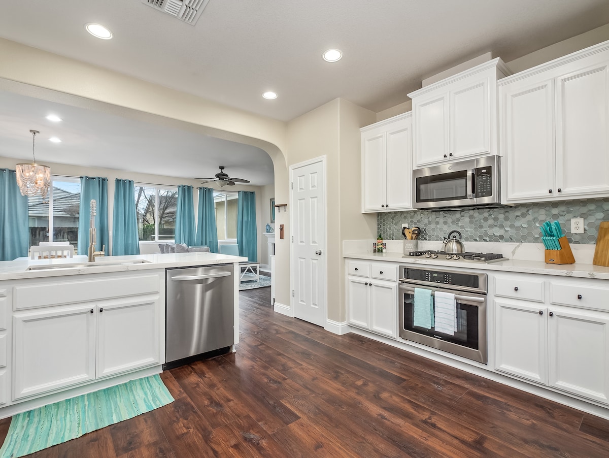 A modern kitchen features white cabinetry and stainless steel appliances. The spacious layout includes a large sink and a stylish backsplash, complemented by warm wood flooring. Bright overhead lights illuminate the area, while natural light filters through nearby curtains, enhancing the inviting space.