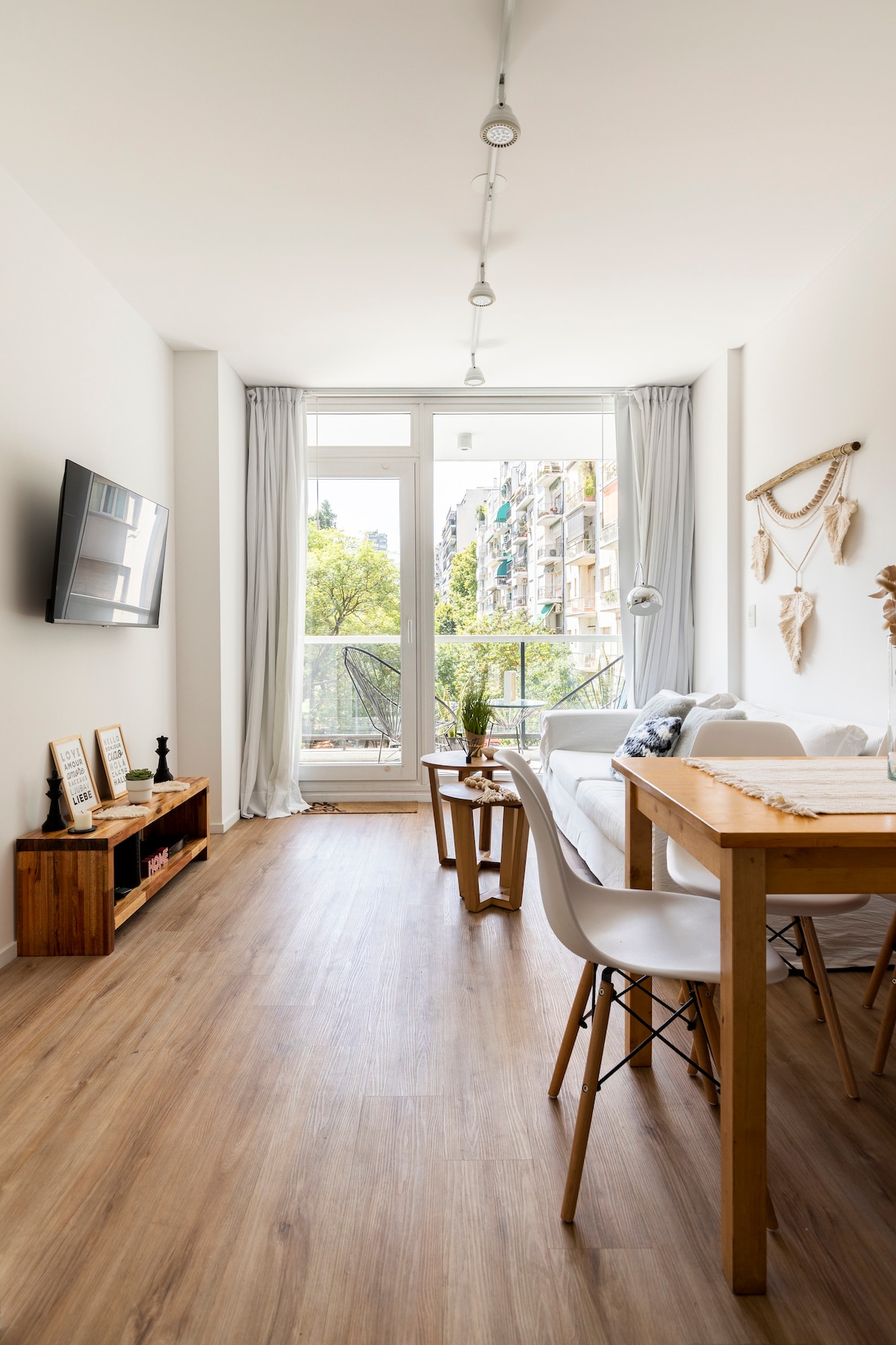 A well-lit living area is showcased with large windows offering a view of greenery outside. A comfortable sofa is arranged alongside a wooden dining table and stylish chairs, with a flat-screen TV mounted on the wall. Light wood flooring adds to the ambiance.