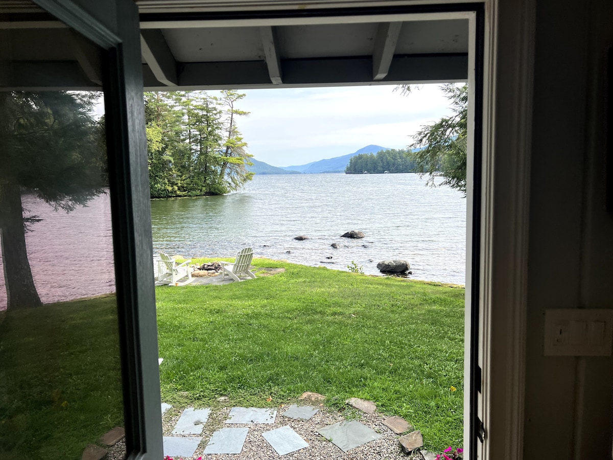 A view of the serene lake is framed by an open door, showcasing a lush green lawn leading to the water. Stone pathways guide the way, while Adirondack chairs are arranged around a fire pit. Trees line the shore, with distant mountains visible across the lake.