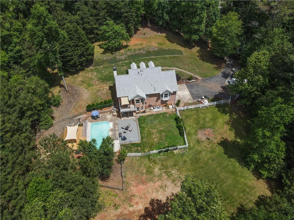 An aerial view captures a spacious home surrounded by trees, featuring a swimming pool and outdoor seating area. The property includes a grassy yard, bordered by a fence, with ample landscaping visible throughout the surroundings.