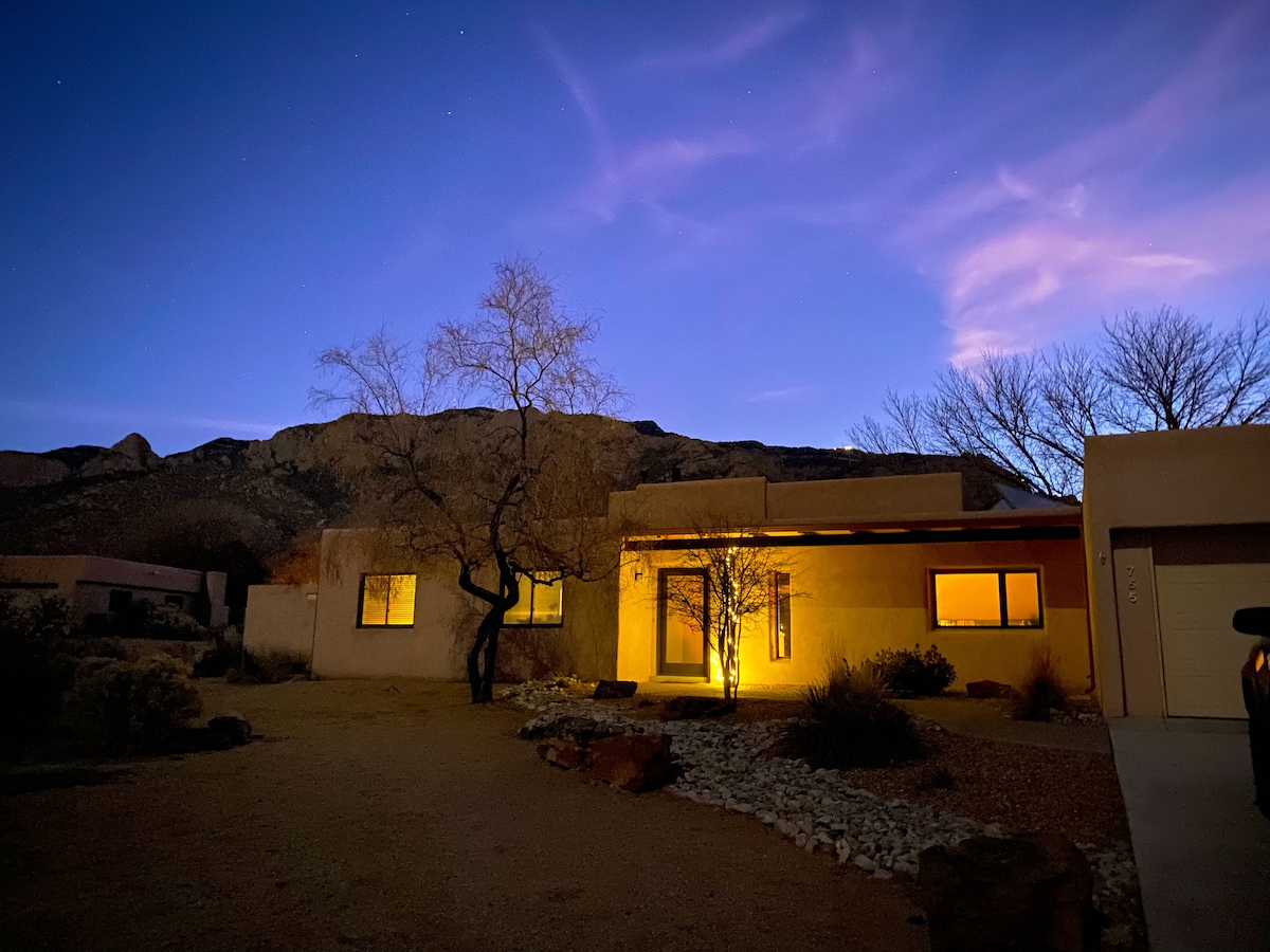 The home is illuminated softly against a twilight sky, with warm light spilling from the windows. A tree stands in the foreground, and the mountain is visible in the background, enhancing the serene desert setting.