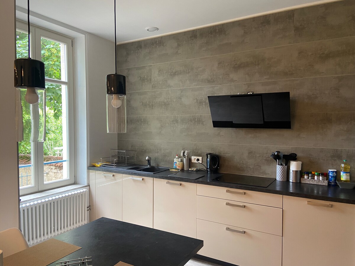 A modern kitchen features sleek cabinetry and a dark countertop. Large windows illuminate the space, bringing in natural light. Pendant lights hang above the central island, complementing the minimalist design. Kitchen appliances and utensils are neatly arranged, contributing to an organized ambiance.