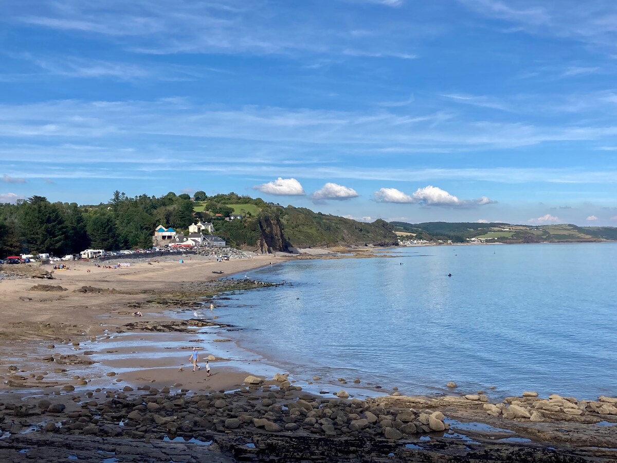 A serene beach scene is captured, showcasing a gentle shoreline. Calm waters reflect the blue sky, and a distant hill is dotted with buildings. Smooth sand is present along the water's edge, with scattered rocks and small groups of people enjoying the area.
