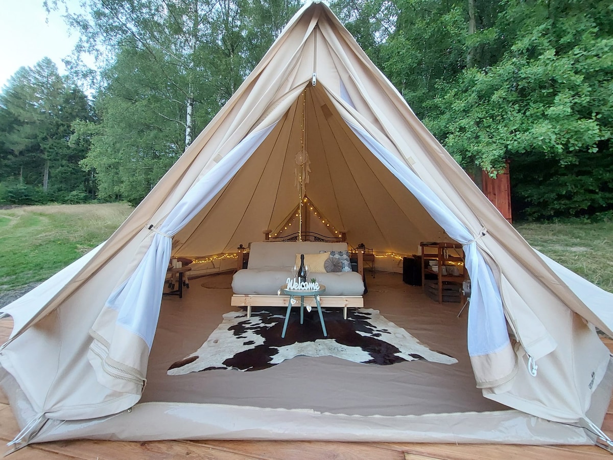 A glamping tent is showcased, featuring a spacious interior with a light-colored bed and simple furnishings. Soft lighting is attached along the tent's structure, creating a gentle ambiance. A patterned rug is positioned on the floor, complementing the natural setting of trees and grass outside.