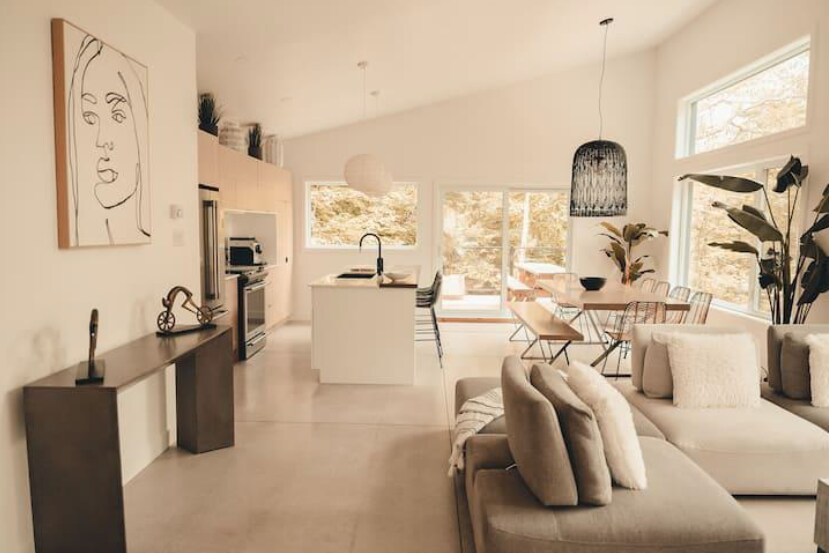 An open living space showcases a modern kitchen with light wood cabinetry and a central island. Large windows provide natural light, revealing views of the outdoors. A comfortable sectional sofa and a sculptural wall piece add interest to the area, complemented by indoor plants.