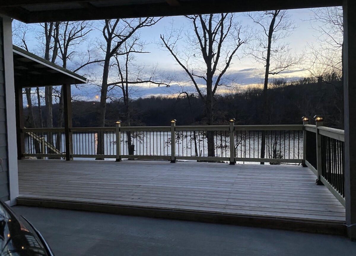 A spacious wooden deck overlooks a tranquil body of water, framed by leafless trees. Soft lighting fixtures are visible along the deck's railing, enhancing the outdoor space, while a partially cloudy sky adds a serene backdrop to the scene.