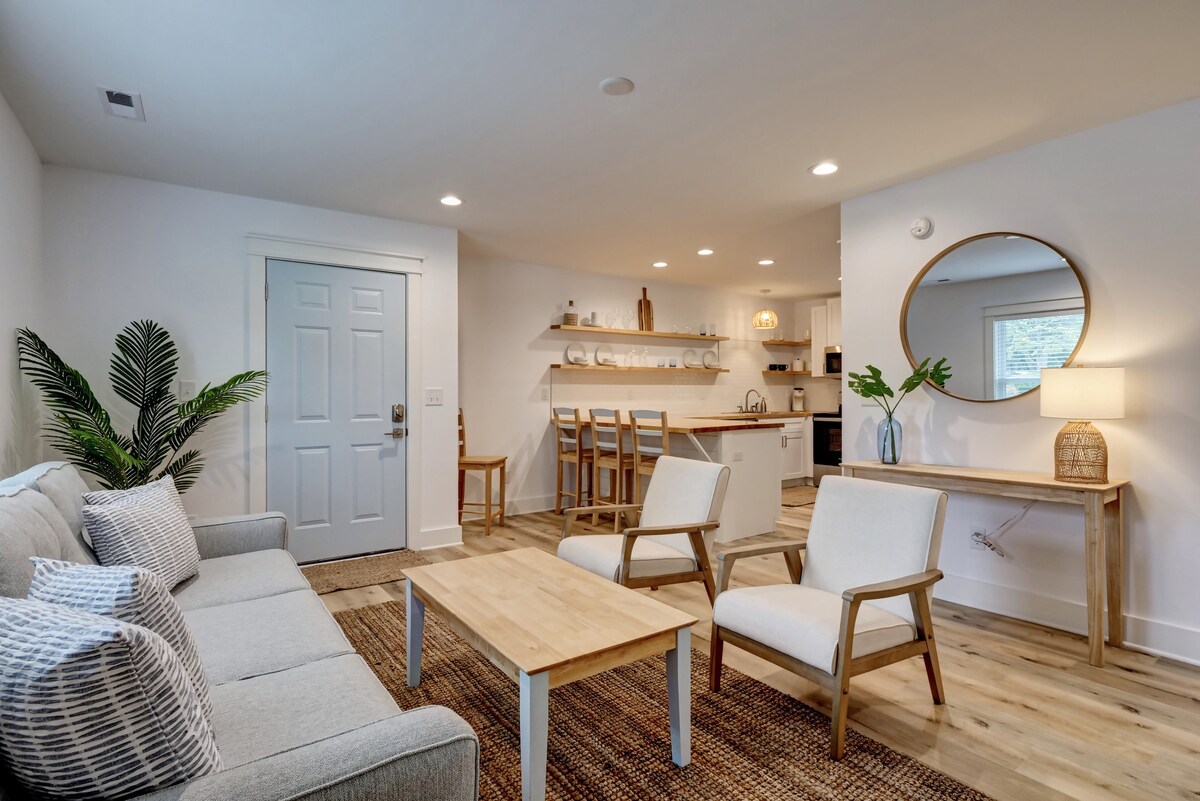 A light-filled living area features a comfortable gray sofa and two wooden chairs surrounding a simple coffee table. The space is accentuated by a round mirror and natural textiles, with an open kitchen area visible in the background, showcasing modern cabinetry and decor.