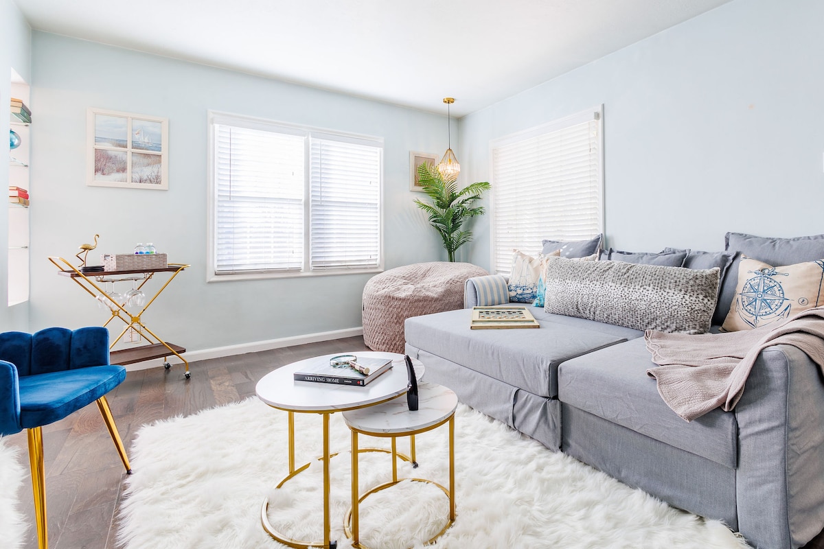 A light-filled living area features a spacious gray sofa, accented with decorative pillows. Two round coffee tables with gold legs rest on a soft white rug. A blue chair is positioned beside a small side table, while a plant adds a touch of greenery by the window.