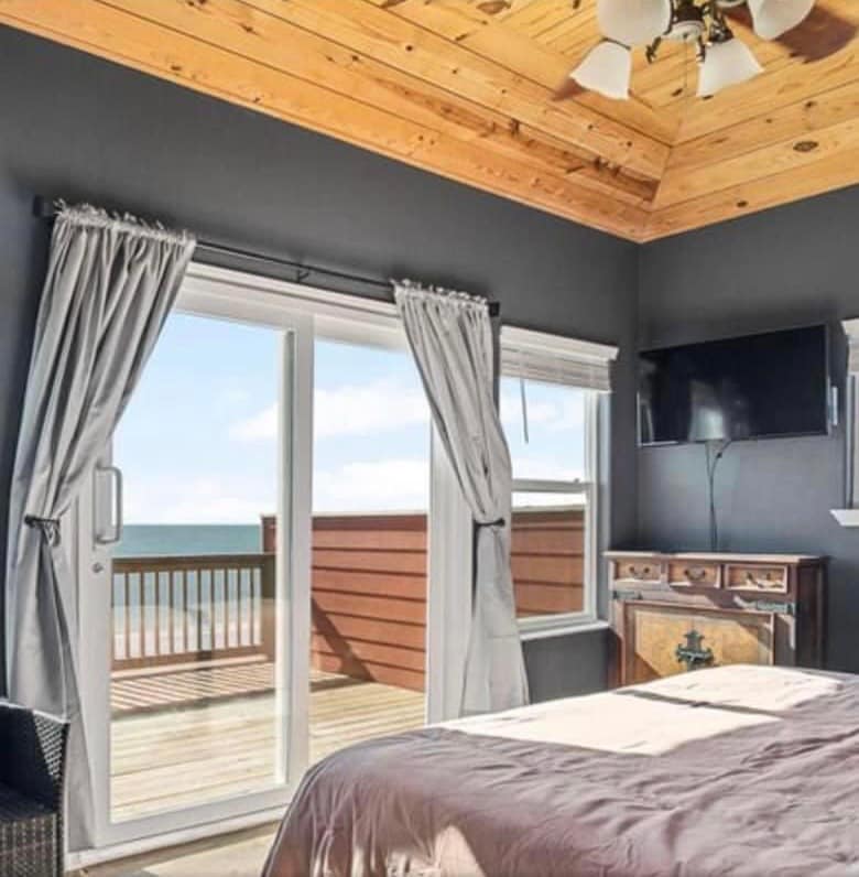 A serene bedroom features a large sliding door that opens to a private balcony with ocean views. The room is adorned with a wood-paneled ceiling and has a wall-mounted television. Gray curtains frame the windows, complementing the dark walls.