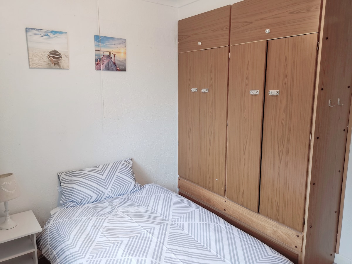 A single bedroom is presented with a neatly made bed adorned with a patterned blanket. A wooden wardrobe occupies one wall, while two small photographs of serene landscapes are displayed above. A side table completes the cozy space.