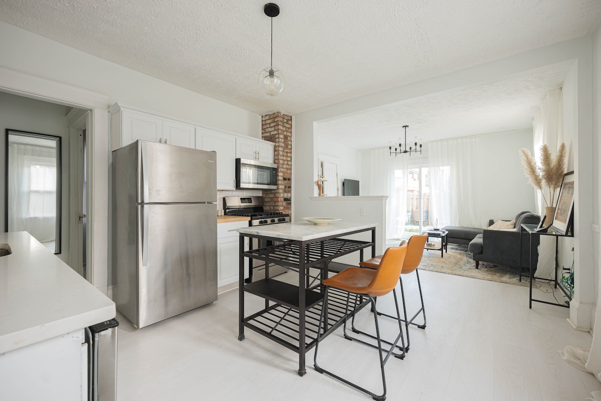 The open kitchen features stainless steel appliances, including a fridge and stove, paired with a central black island. Two orange chairs are positioned at the island, while bright light enhances the spacious interior, leading to a cozy living area with a white sofa and large windows.