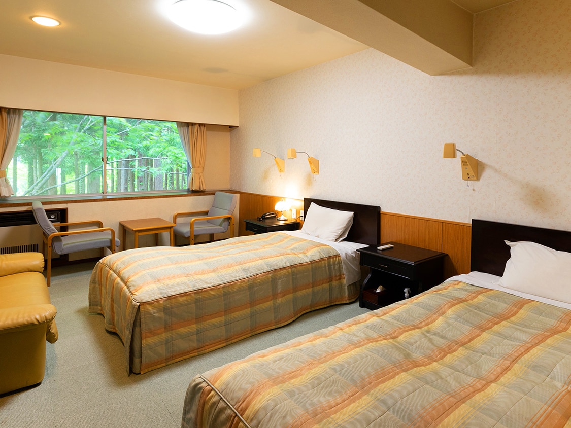 A bright twin room features two spacious beds dressed in light-colored bedding. Two armchairs and small tables are arranged near the window, offering views of greenery. Soft lighting and simple decor create a calm and comfortable environment.