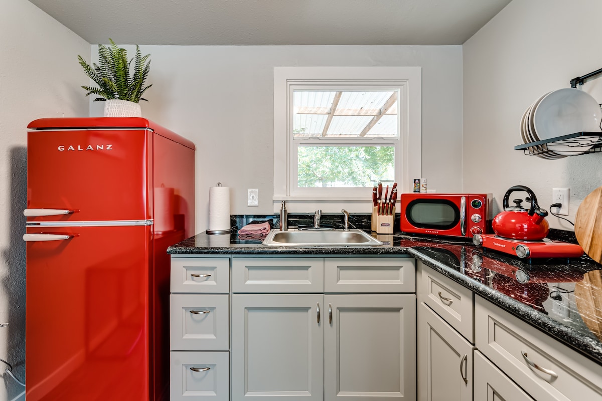 A compact kitchen features modern appliances including a vibrant red refrigerator, microwave, and kettle. Counter space is adorned with kitchen utensils and a cutting board. A sink sits beneath a window, allowing natural light to brighten the area, complemented by subtle green plants.