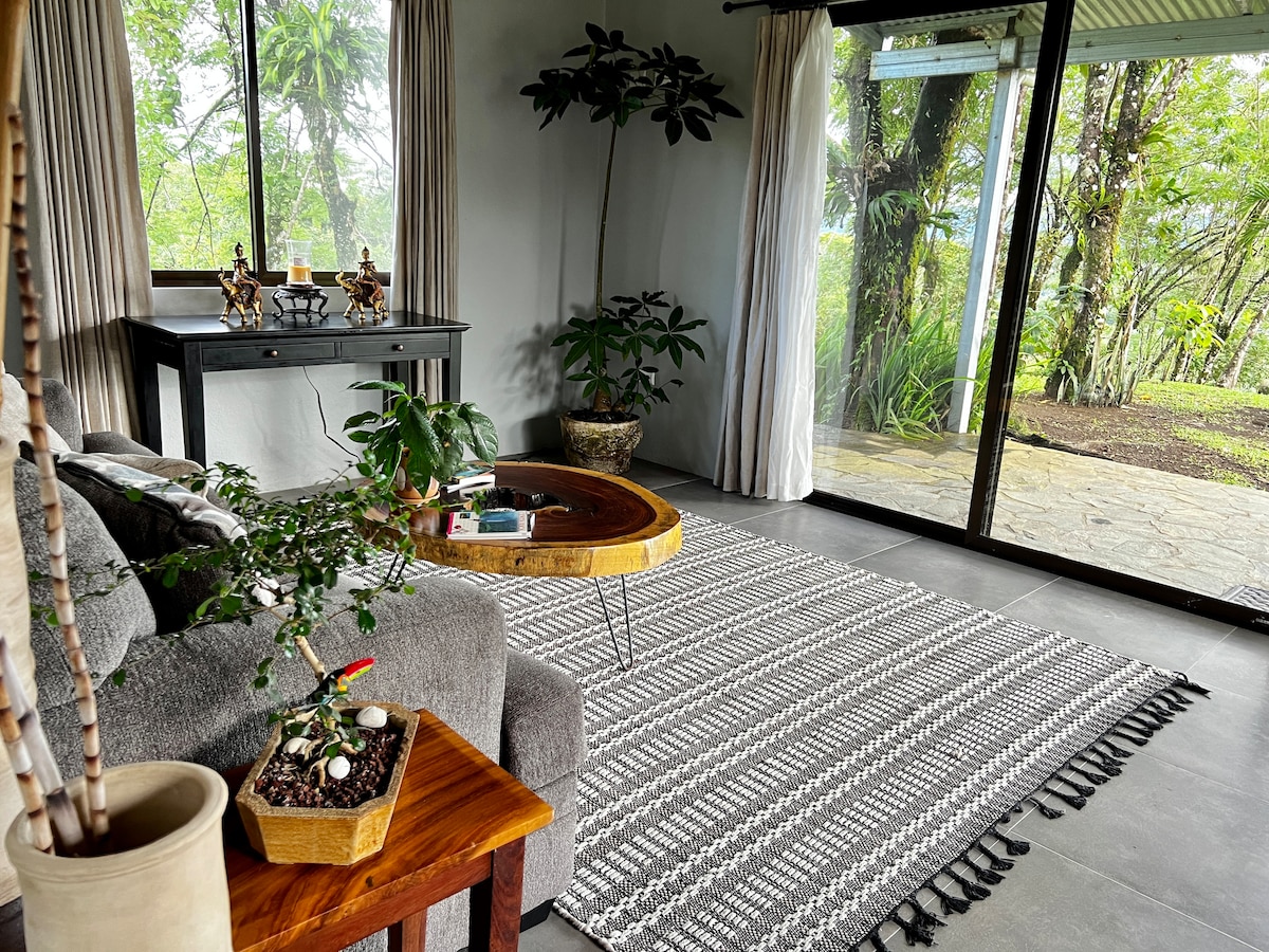 A living area is presented with a comfortable gray sofa and a unique wooden coffee table. A patterned rug covers the floor, complemented by potted plants that add greenery. Large glass doors invite natural light and offer views of the surrounding landscape.