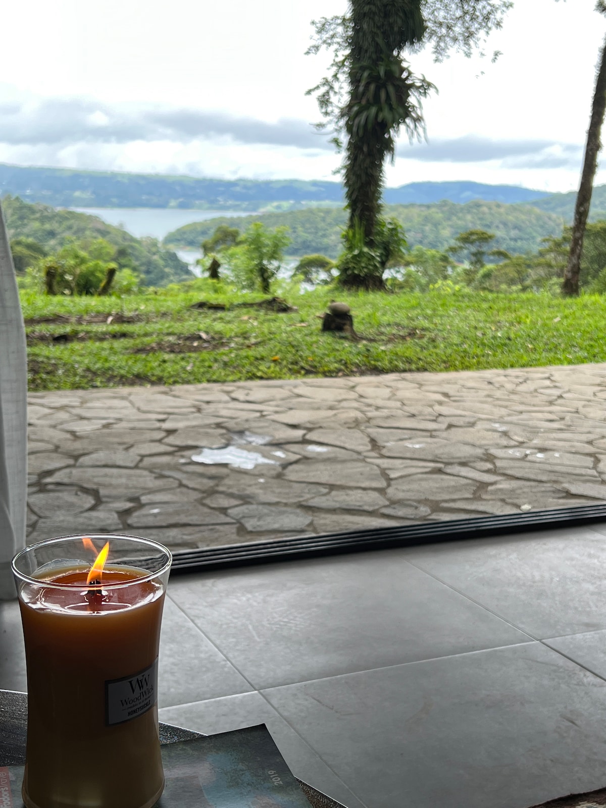 A lit candle is positioned on a table, with a view of Lake Arenal visible through a large glass door. The landscape features lush greenery and distant hills under a partly cloudy sky, creating a serene and calming atmosphere.