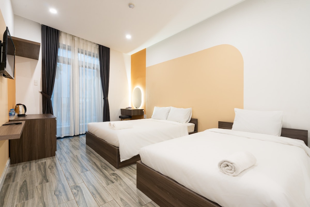 A clean and modern room features two spacious beds and a sleek wooden desk with a television mounted above. Large windows allow natural light to fill the space, complemented by light curtains and an accent wall in a warm shade.