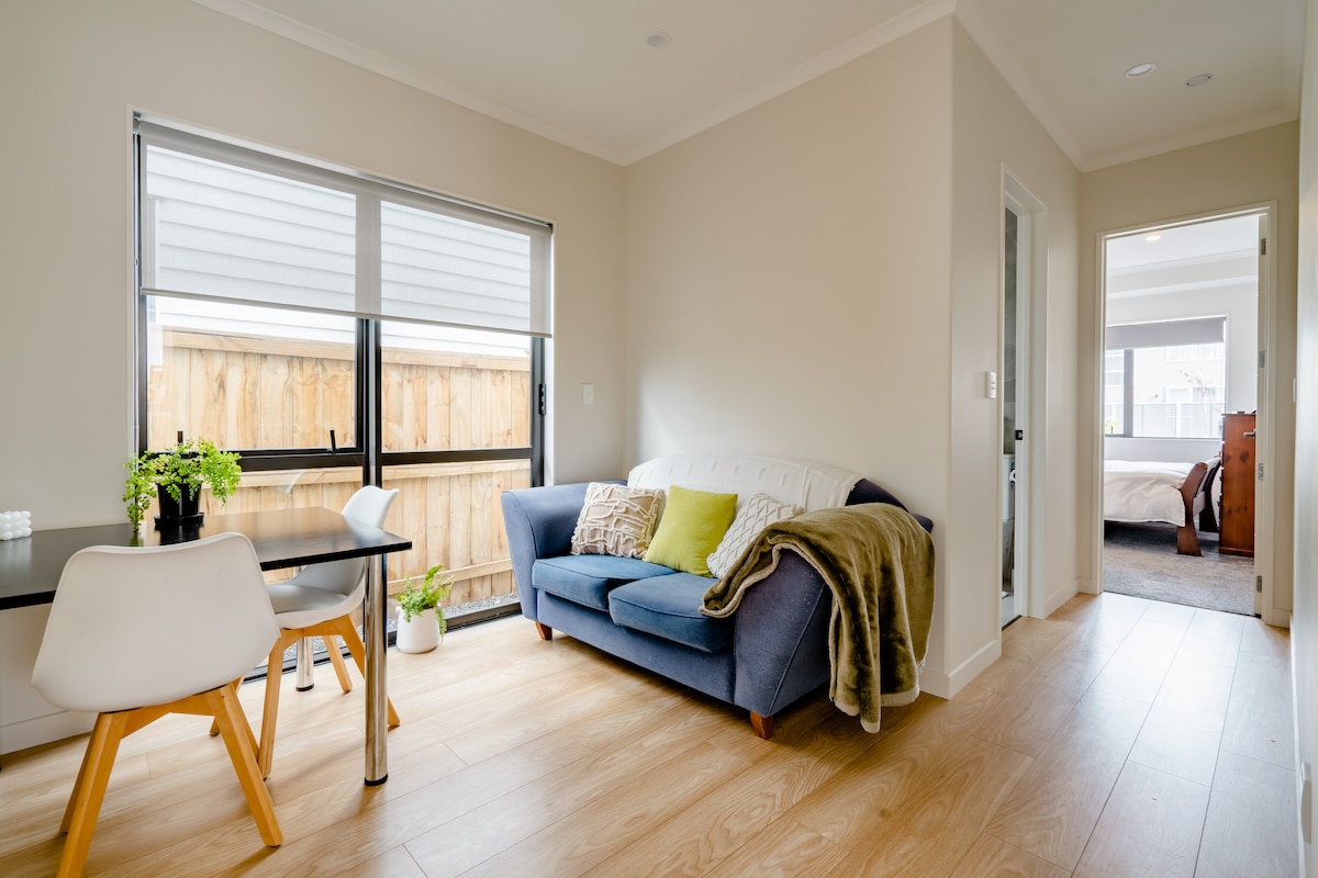 A light-filled lounge area features a comfortable blue sofa adorned with a cozy blanket. A dining table is positioned nearby, complemented by greenery. Natural wood flooring extends throughout the space, with large windows offering views of the outdoor area.