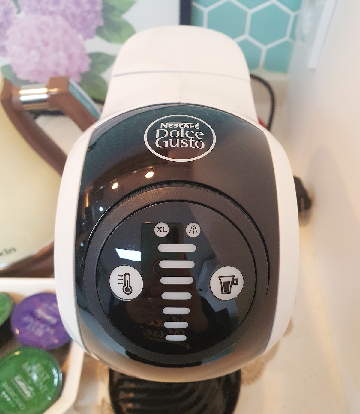 A Nesca fé Dolce Gusto coffee machine is displayed on a kitchen countertop. The machine features an intuitive control panel with options for different beverage sizes. Surrounding it are coffee pods in various colors, creating a functional coffee-making setup.