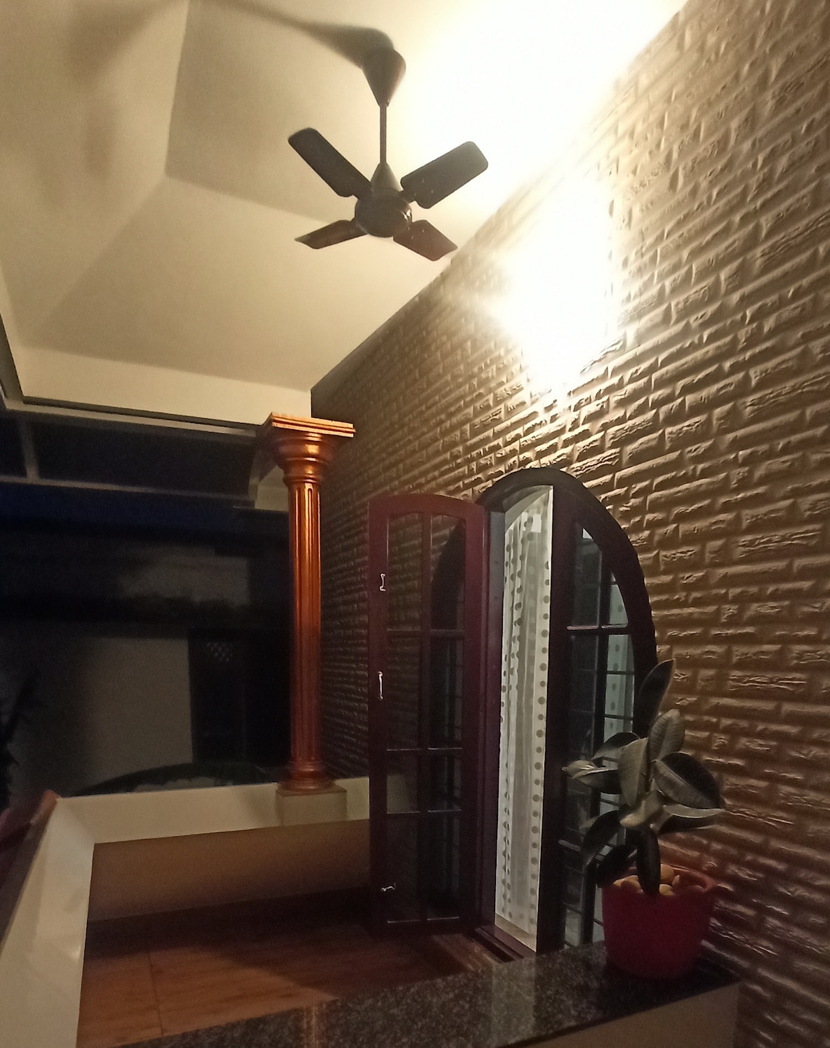 A balcony area is shown with warm lighting, featuring a ceiling fan and decorative elements. A patterned wall and a plant in a pot enhance the inviting space. The double window frame allows for natural light and views, contributing to a cozy, open atmosphere.