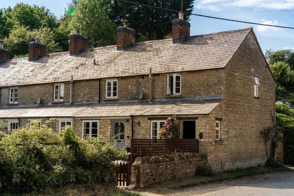 The cottage features a warm stone exterior, blending harmoniously with the surrounding greenery. A wooden fenced area is visible in front, enhancing the cottage's charm. Multiple windows allow natural light to fill the interior, while a welcoming entrance invites guests inside.