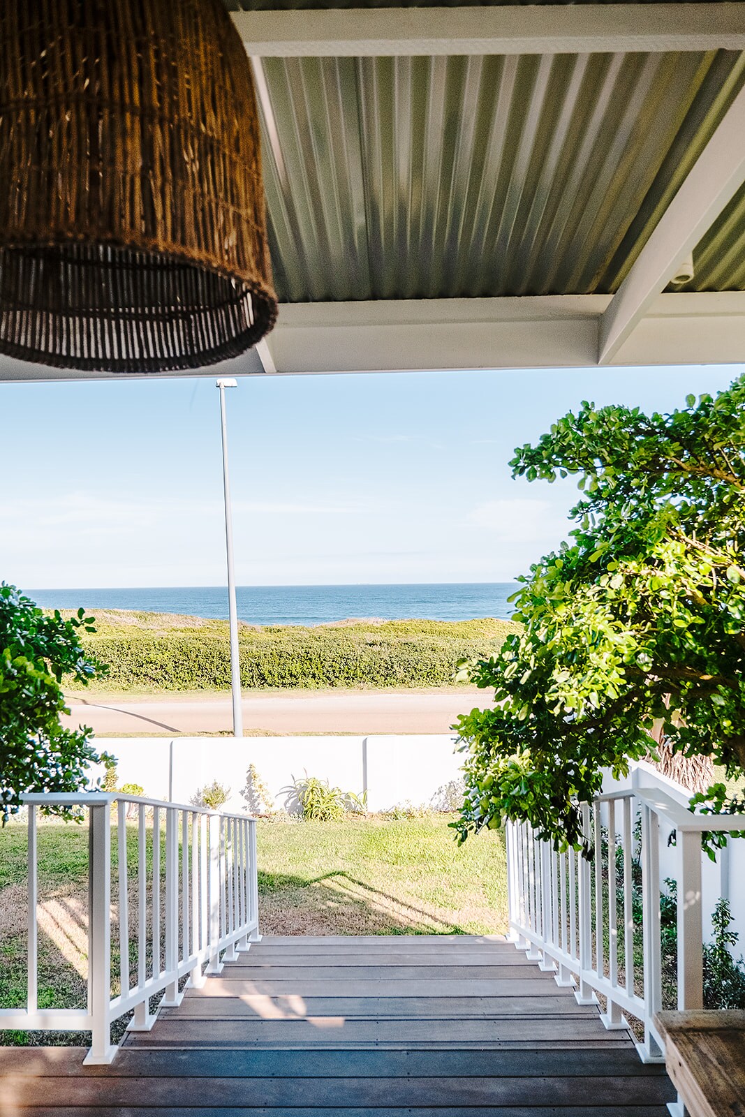 A view from a private deck showcases a clear seascape, framed by lush greenery on either side. Steps lead down towards a grassy area, with a road visible in the foreground and calm waters beyond.